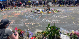 Individuals circled around a memorial in the street for George Floyd. There are drawings on the ground written in chalk and flowers in the center.  