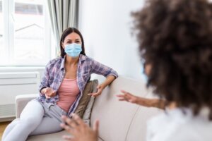 Two women wearing masks talk on a couch in a sunlit room