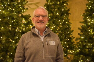 Tom Olson standing in front of three Christmas trees at the congregation convening. Headshot. 
